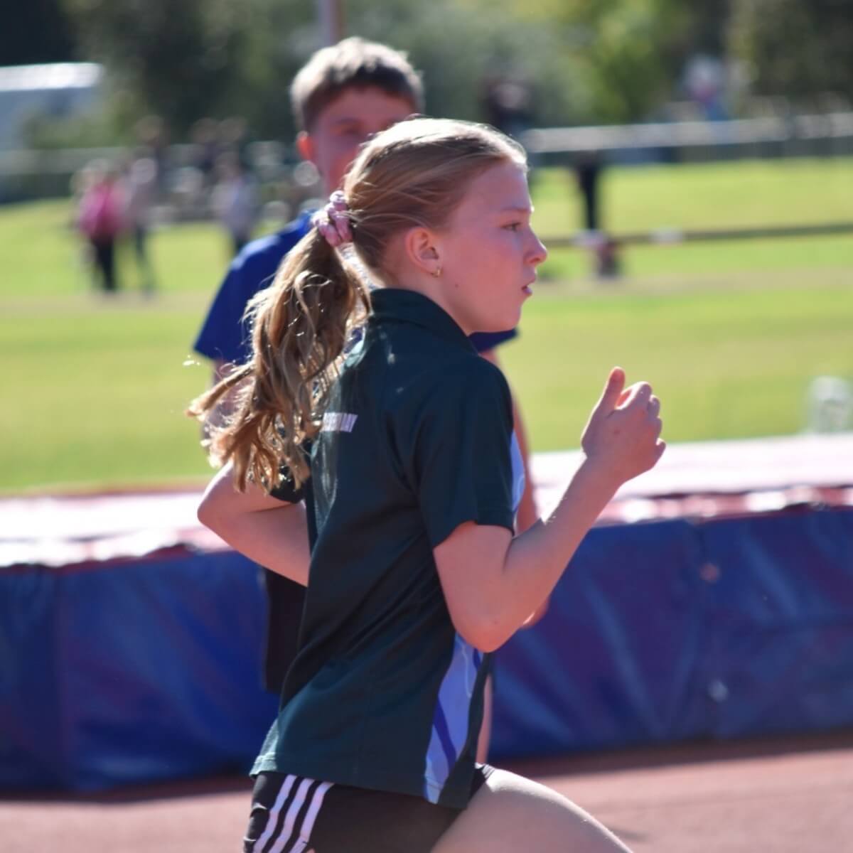 A young girl with long hair runs on a track, wearing a dark shirt and athletic shorts, with another child in the background.