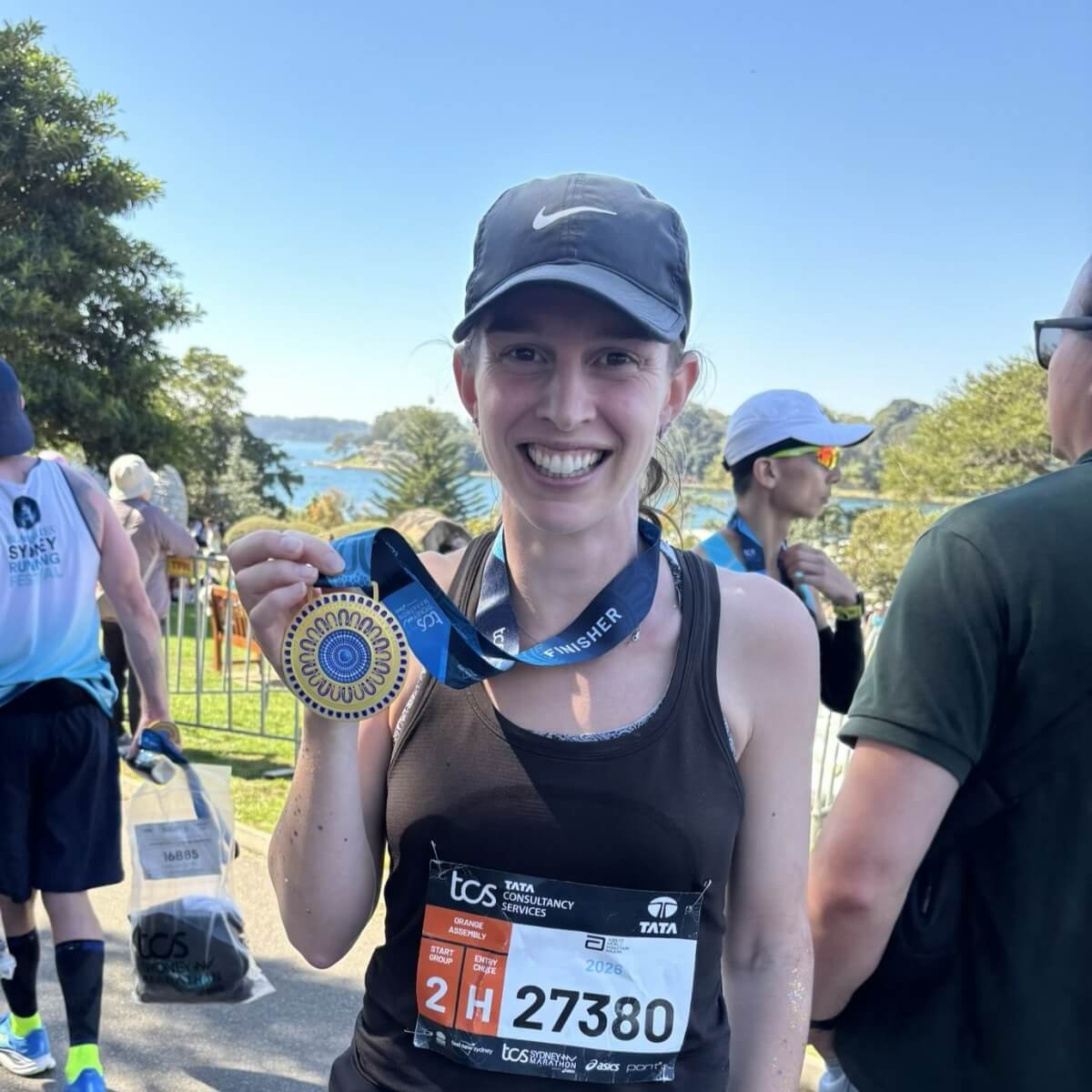 A smiling woman in a black tank top holds a marathon medal, with a scenic background and other runners nearby.