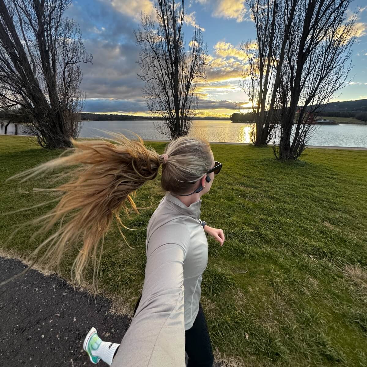 A woman runs along a path by a lake at sunset, with trees and grass in the background.