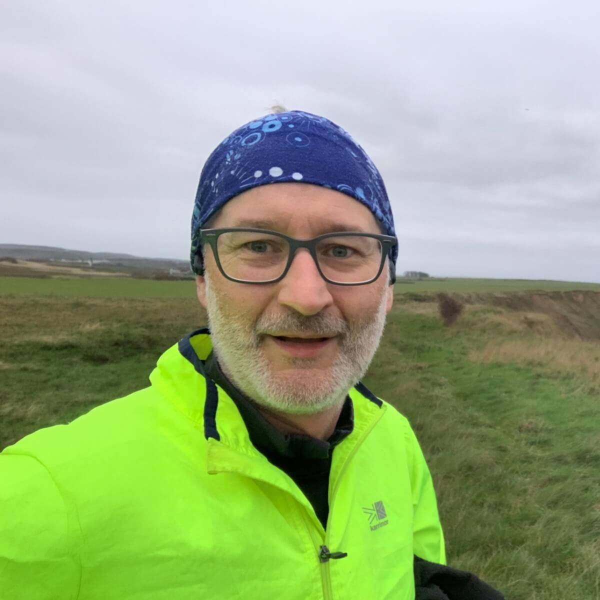 A man in a bright yellow jacket and blue headband smiles outdoors, with grassy fields and a cloudy sky in the background.