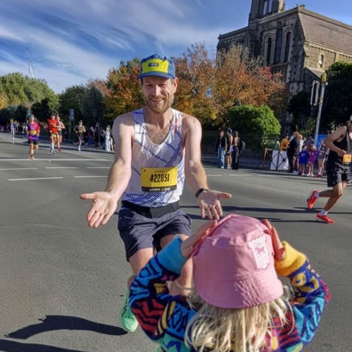 A runner in a tank top reaches out to a child wearing a colorful sweater and pink hat during a marathon.