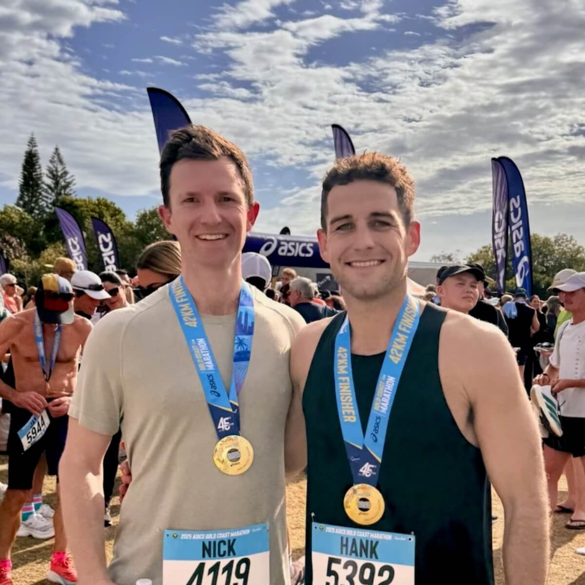 Two men stand smiling with medals around their necks at a marathon event, with spectators and flags in the background.
