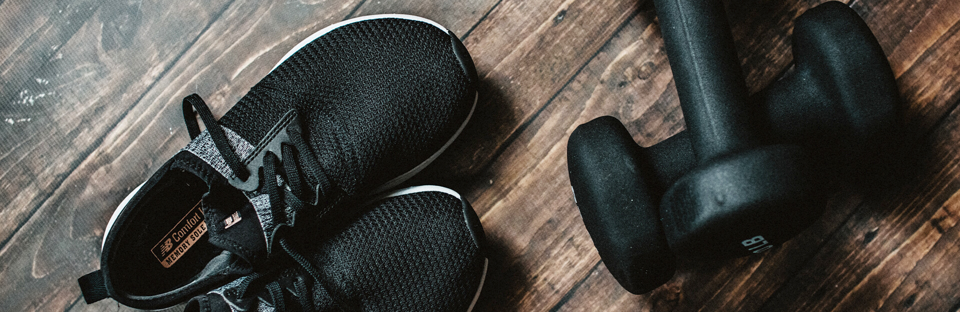 A pair of black athletic shoes and a black dumbbell on a wooden floor.