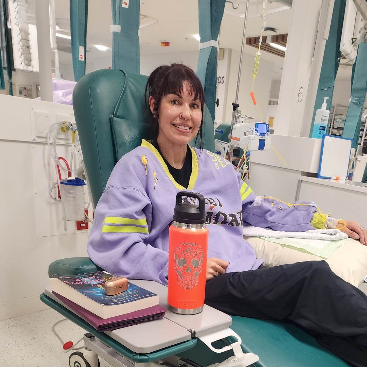 A woman sits in a medical chair, smiling, with a water bottle and books on the side table in a hospital setting.