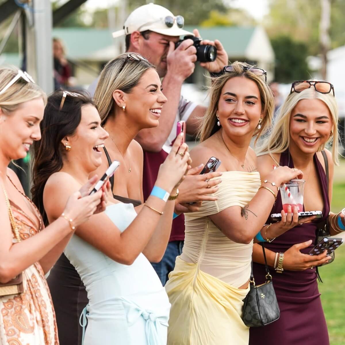 A group of five women, smiling and holding phones, enjoying an outdoor event while a man takes a photo in the background.