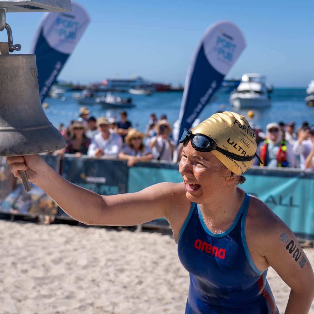 A swimmer in a blue swimsuit and gold cap rings a bell, smiling, with spectators and boats in the background.