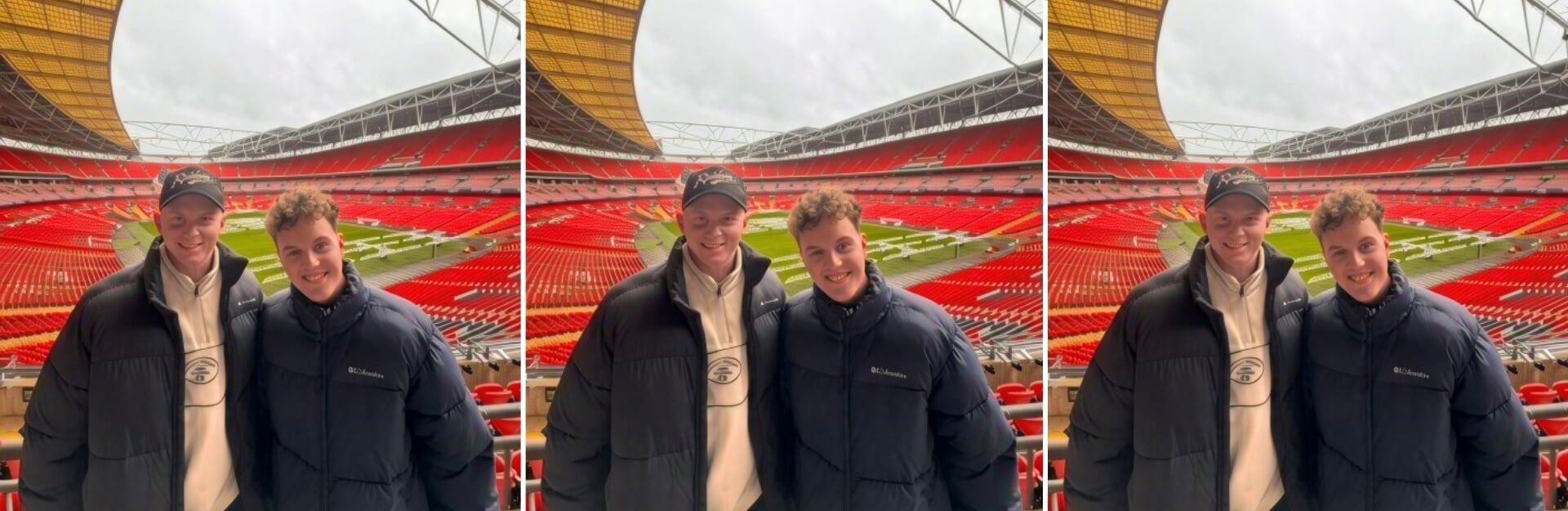 Two young men in puffer jackets smile together in a stadium with red seats and a green field in the background.