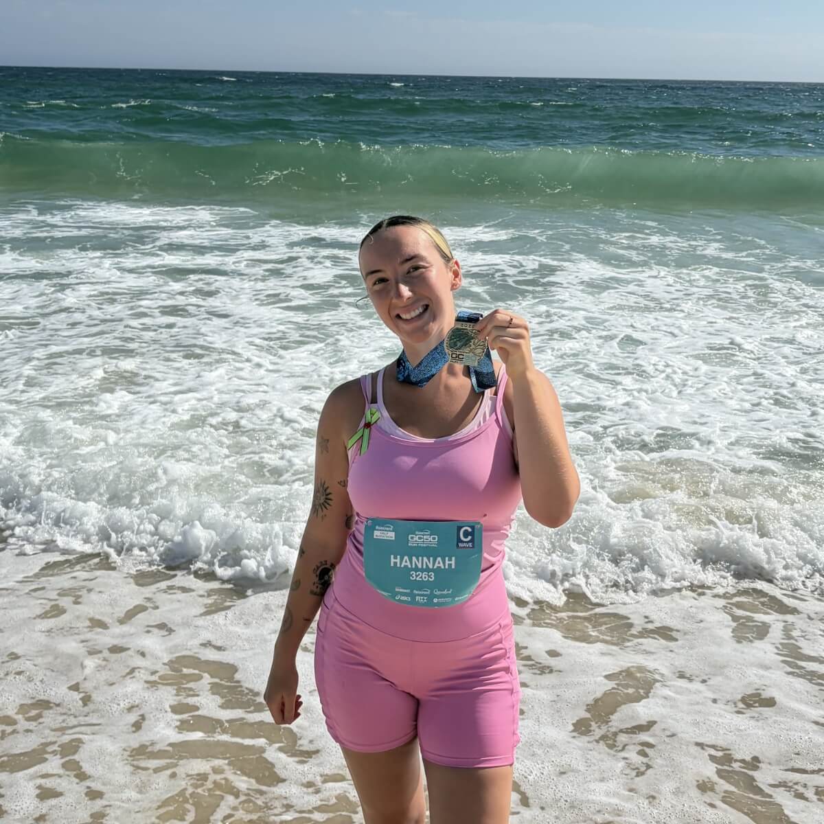 A person in a pink outfit stands on the beach, holding a medal, with waves crashing behind them.