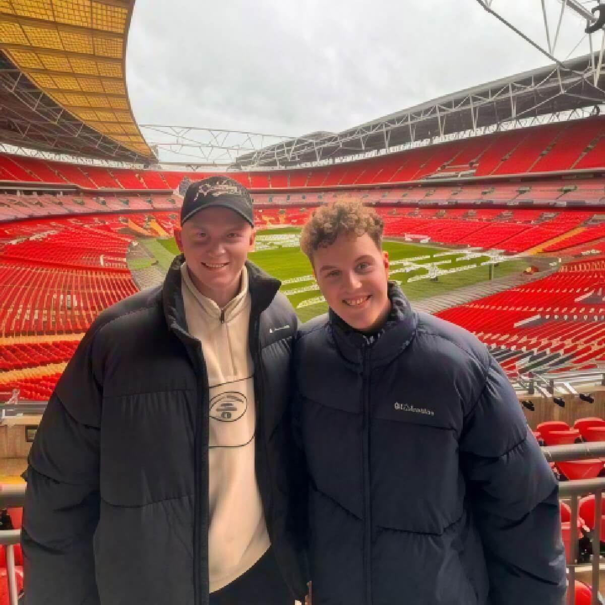 Two young men in puffer jackets smile for a photo at Wembley Stadium, with empty red seats and a green field in the background.